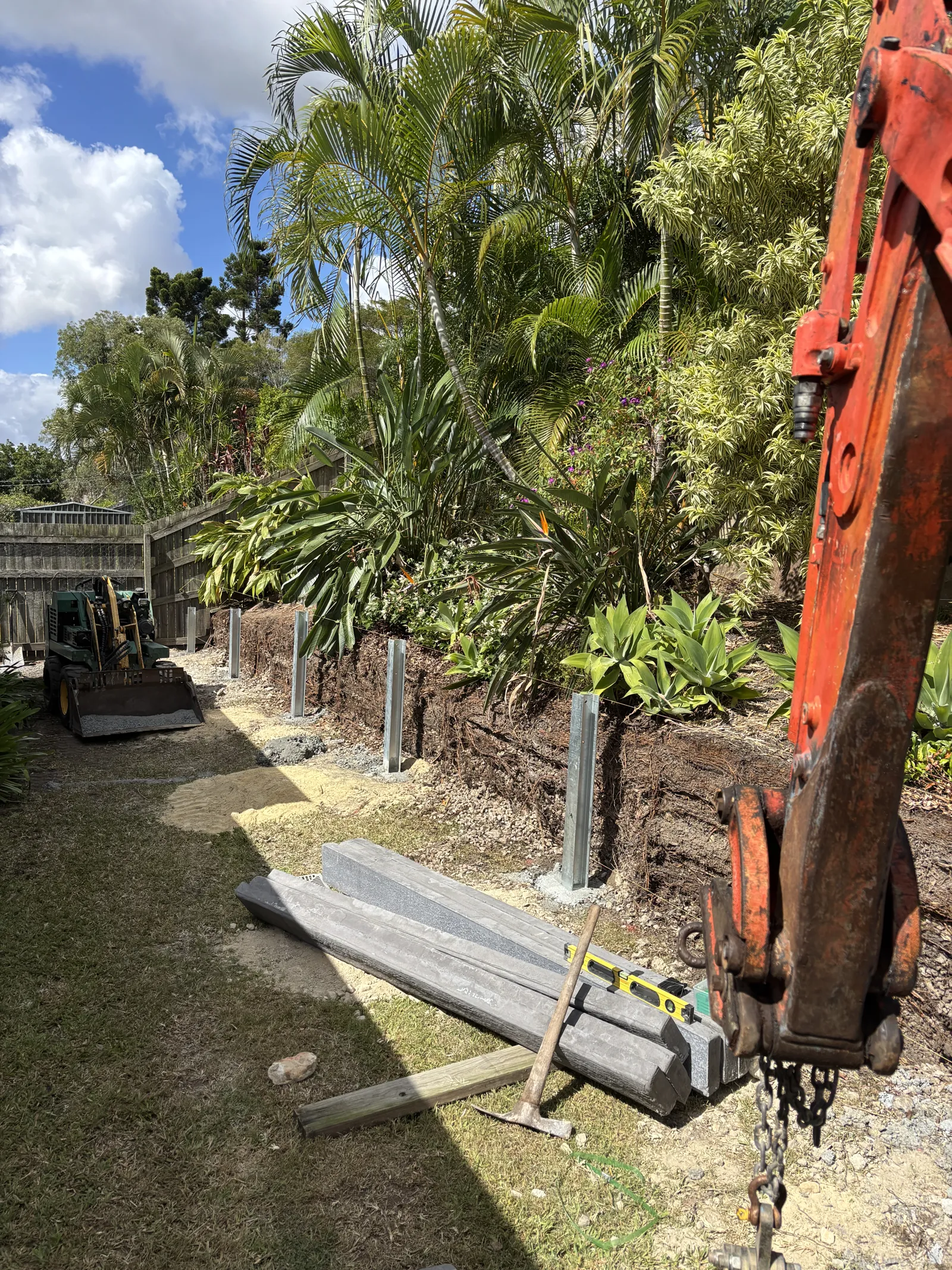 Mini excavator lifting a concrete sleeper into position between galvanised posts